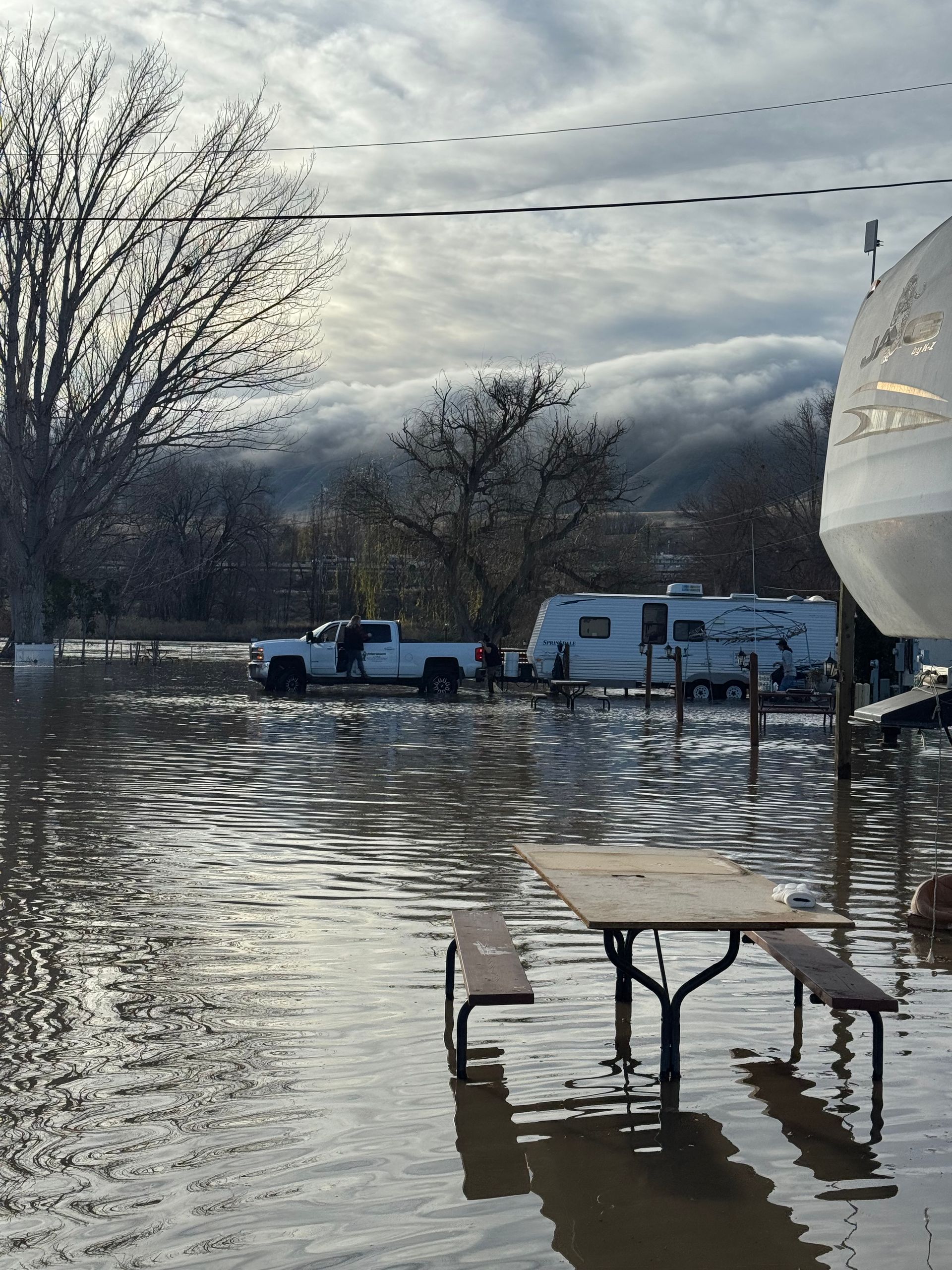 Flooded area with a pickup truck and RV, picnic table submerged. Cloudy sky, trees in the background.