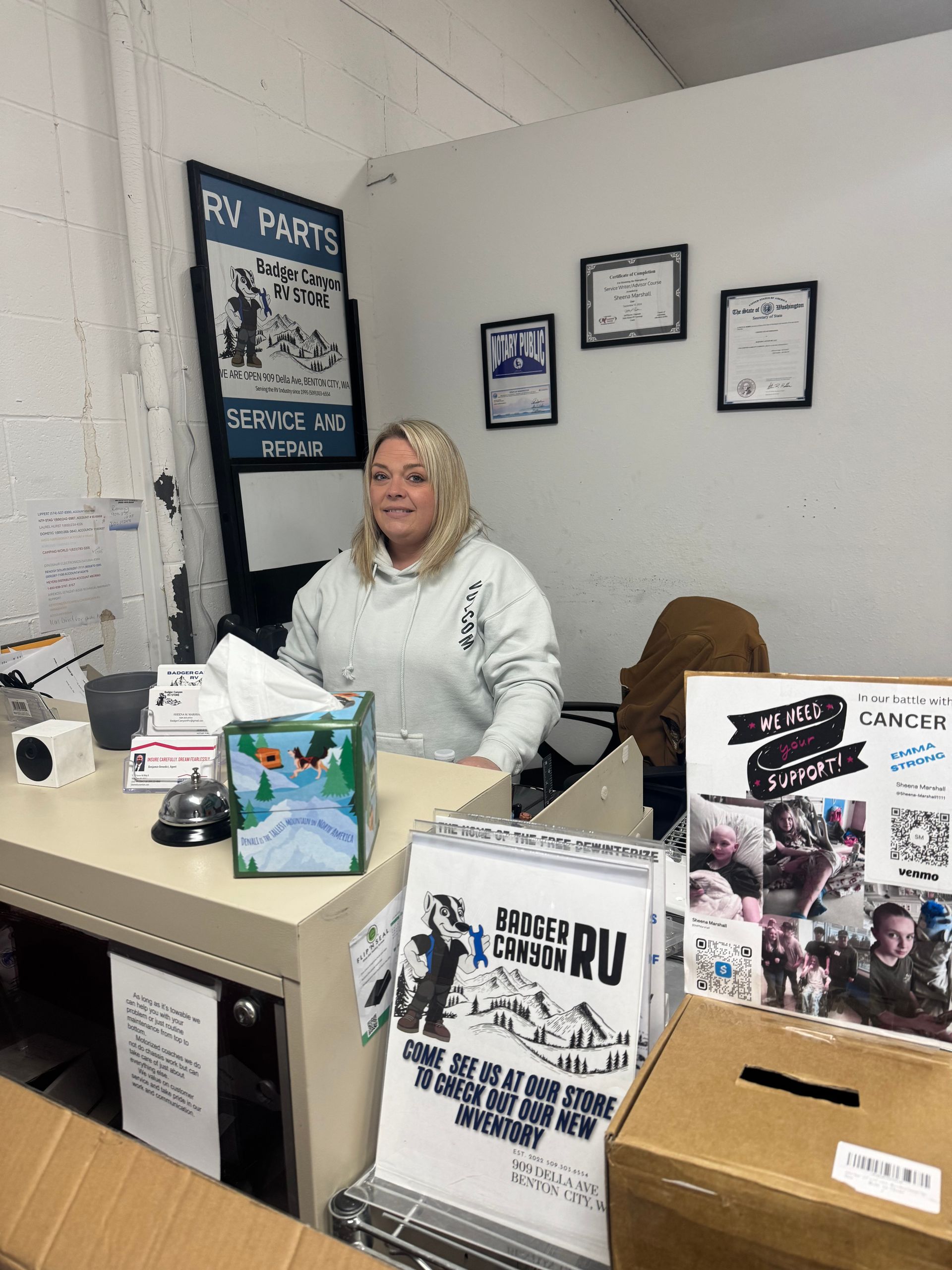 Woman at a desk, smiling. Behind her are posters and framed documents. Boxes and a tissue box are on the desk.