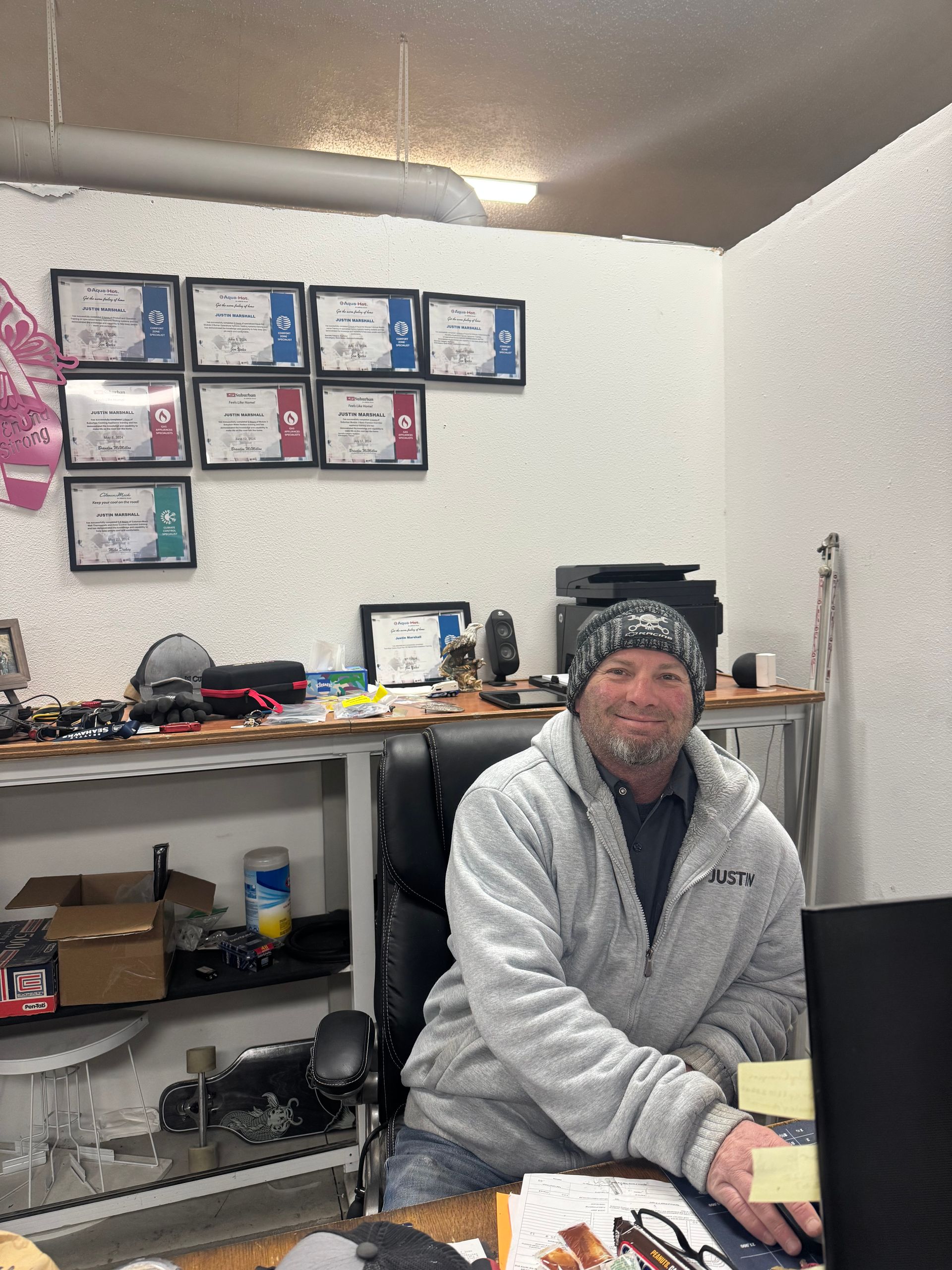Man wearing a beanie sits at a desk in an office. Certificates are on the wall behind him.