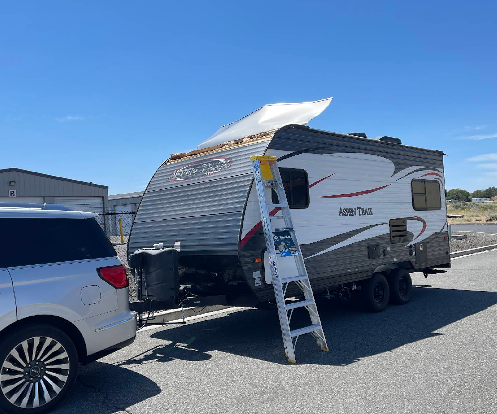 Damaged RV with exposed roof, parked next to a silver SUV on a sunny day. A ladder leans against the RV.