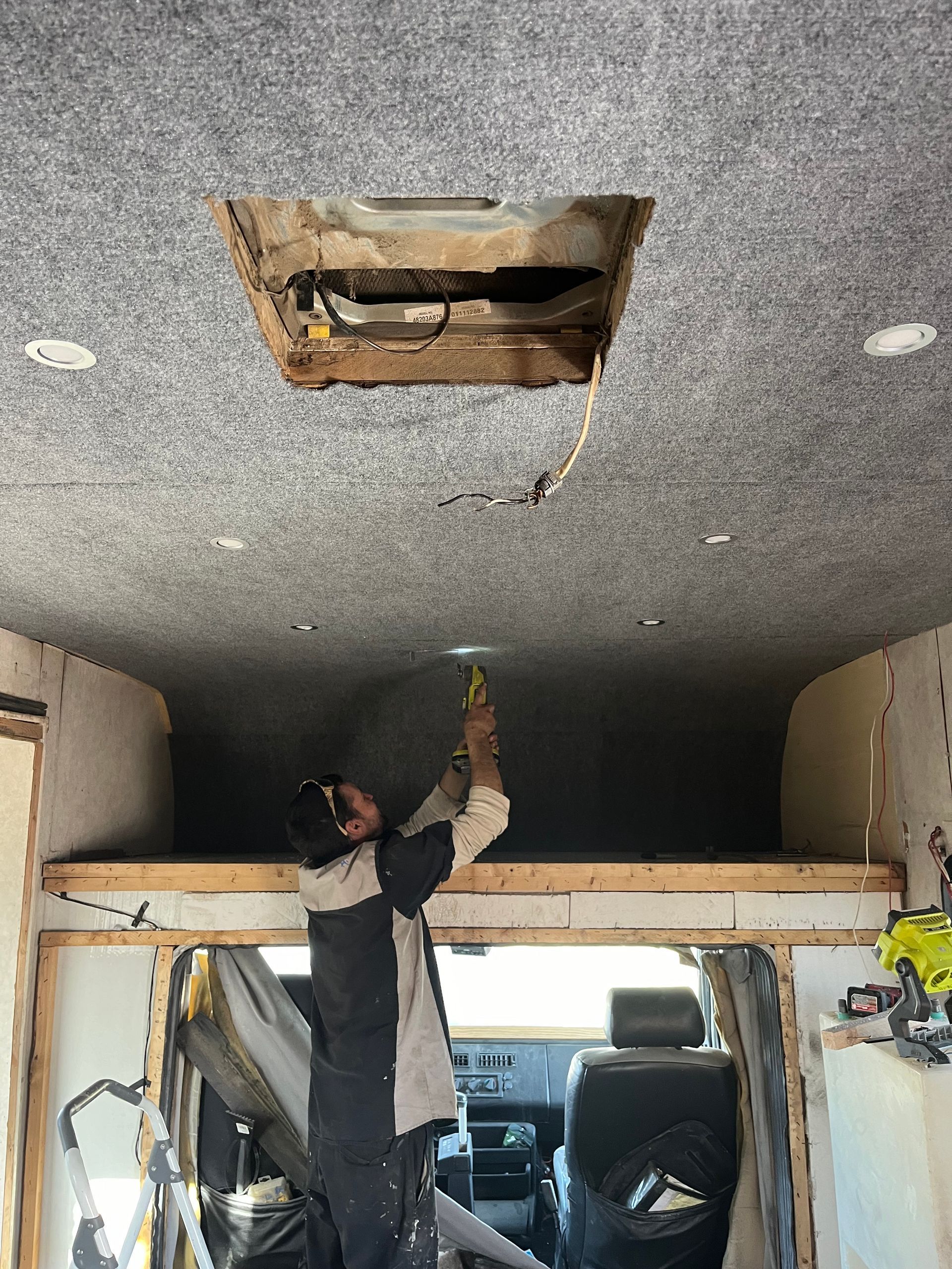 Person works on a van ceiling, removing a panel. Interior view showing bed frame and tools.