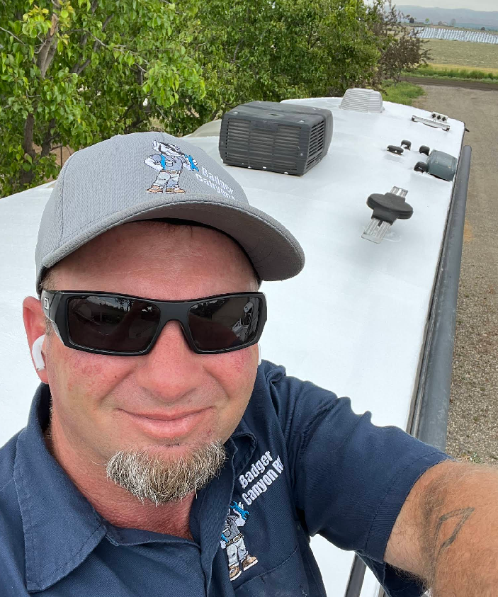 Man wearing sunglasses and a cap on a RV roof, smiling at the camera. White roof with vents.