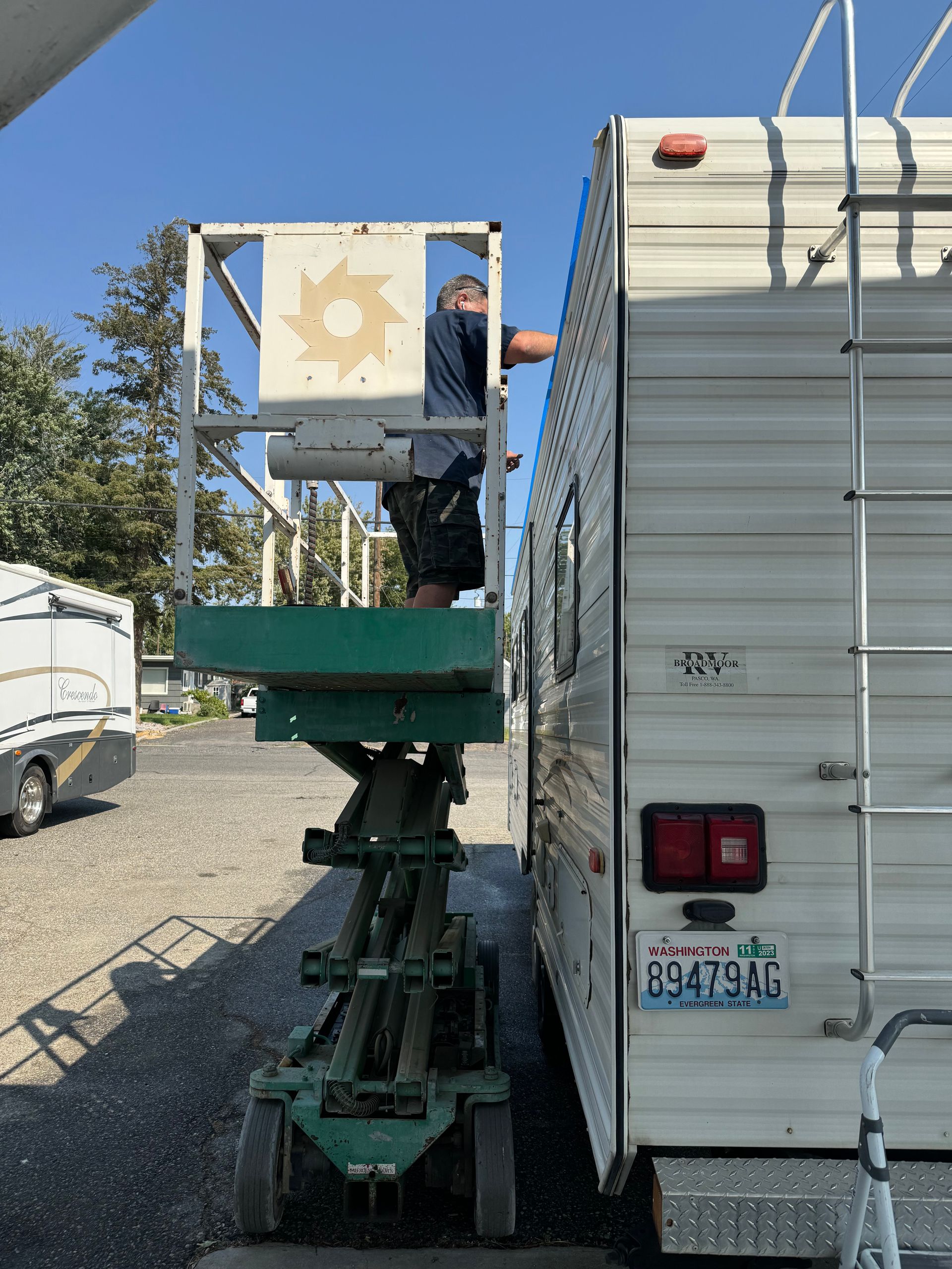 Person in a green lift working on the side of a white RV. Sunny outdoor setting.