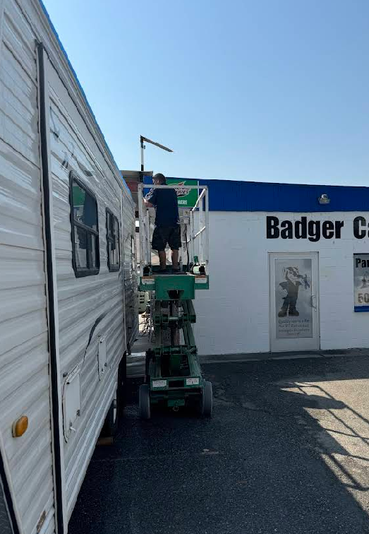 Person on a green lift works on the side of a white RV next to a building labeled 