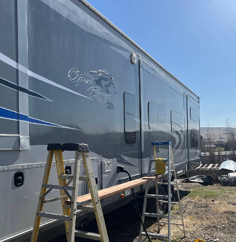 Gray RV with logo and blue stripe, with ladders and plank set up alongside. Sunny outdoor setting.