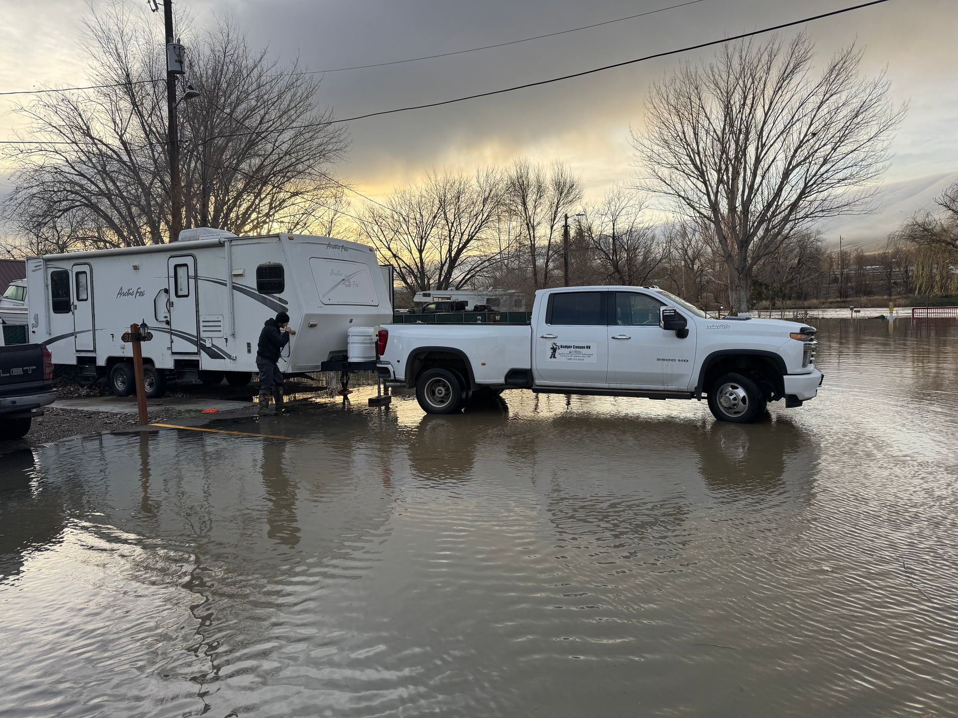 White truck towing camper in floodwater, person near hitch. Cloudy day.