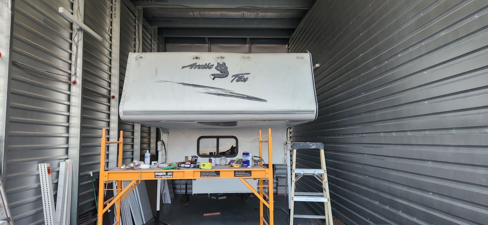 A camper is in a storage unit. It is being worked on with scaffolding and a ladder.