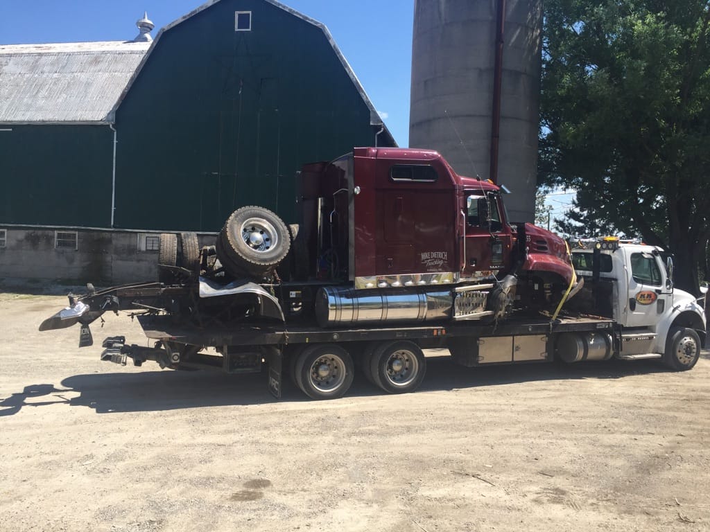 A maroon semi-truck cab on a flatbed tow truck in front of a green barn and silo on a sunny day.