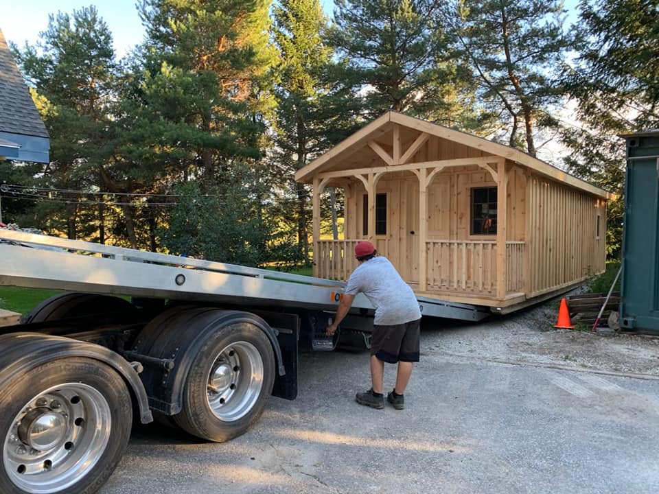 Man guiding a cabin off a flatbed truck. The cabin is wooden, with a small porch and roof. Green trees in the background.