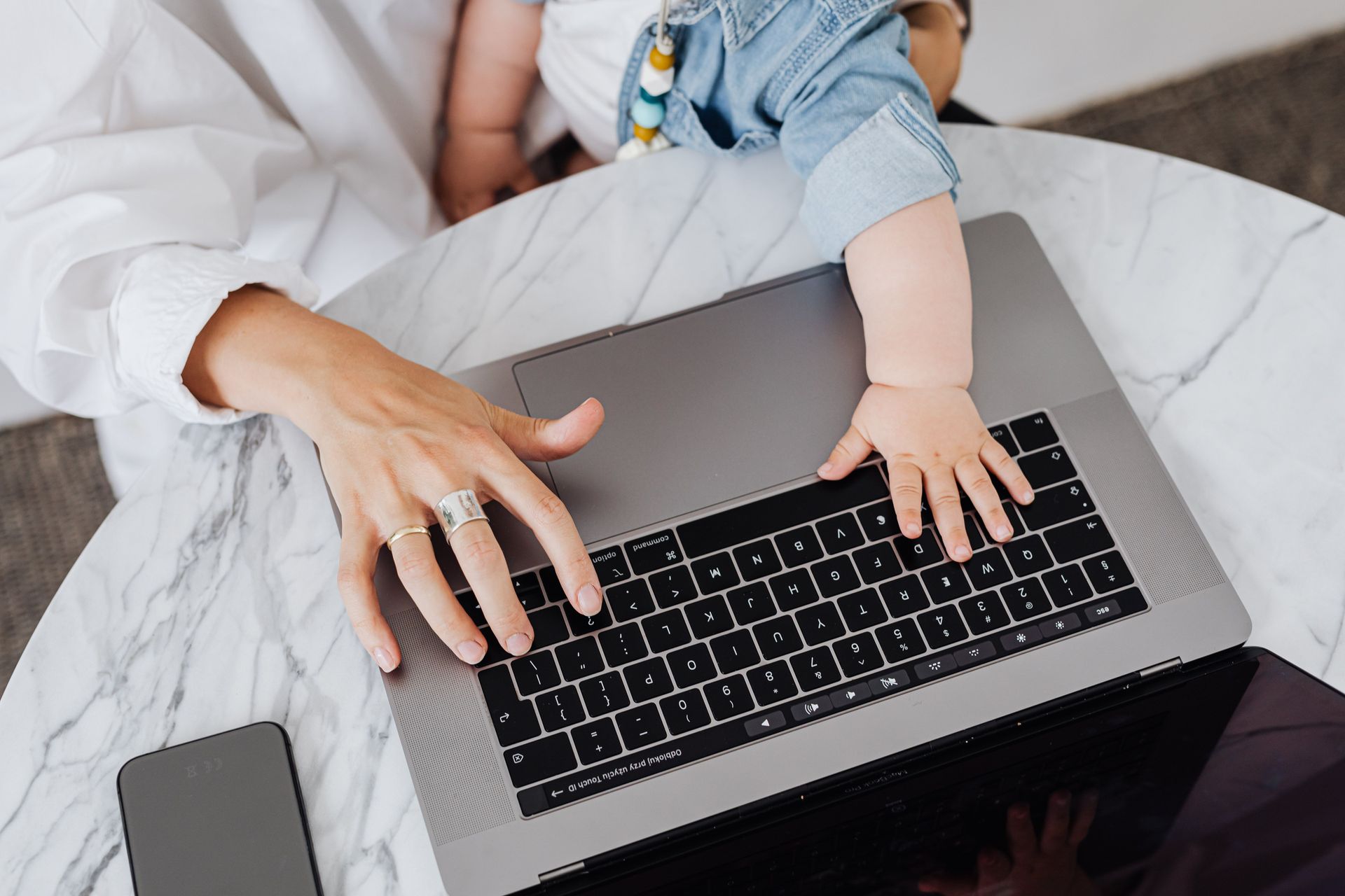 Herausforderung Vereinbarkeit Frau sitzt mit Baby auf dem Schoß an einem Tisch vor einem Laptop und arbeitet.