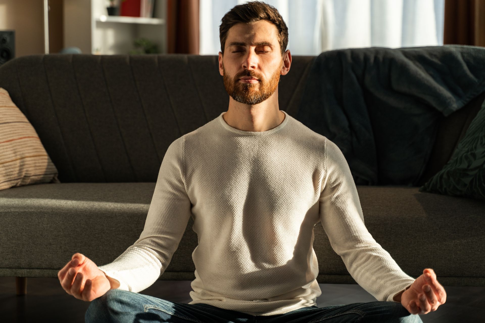Mann sitzt im Schneidersitz von der Sonne angestrahlt auf dem Boden vor seinem Sofa & ist entspannt