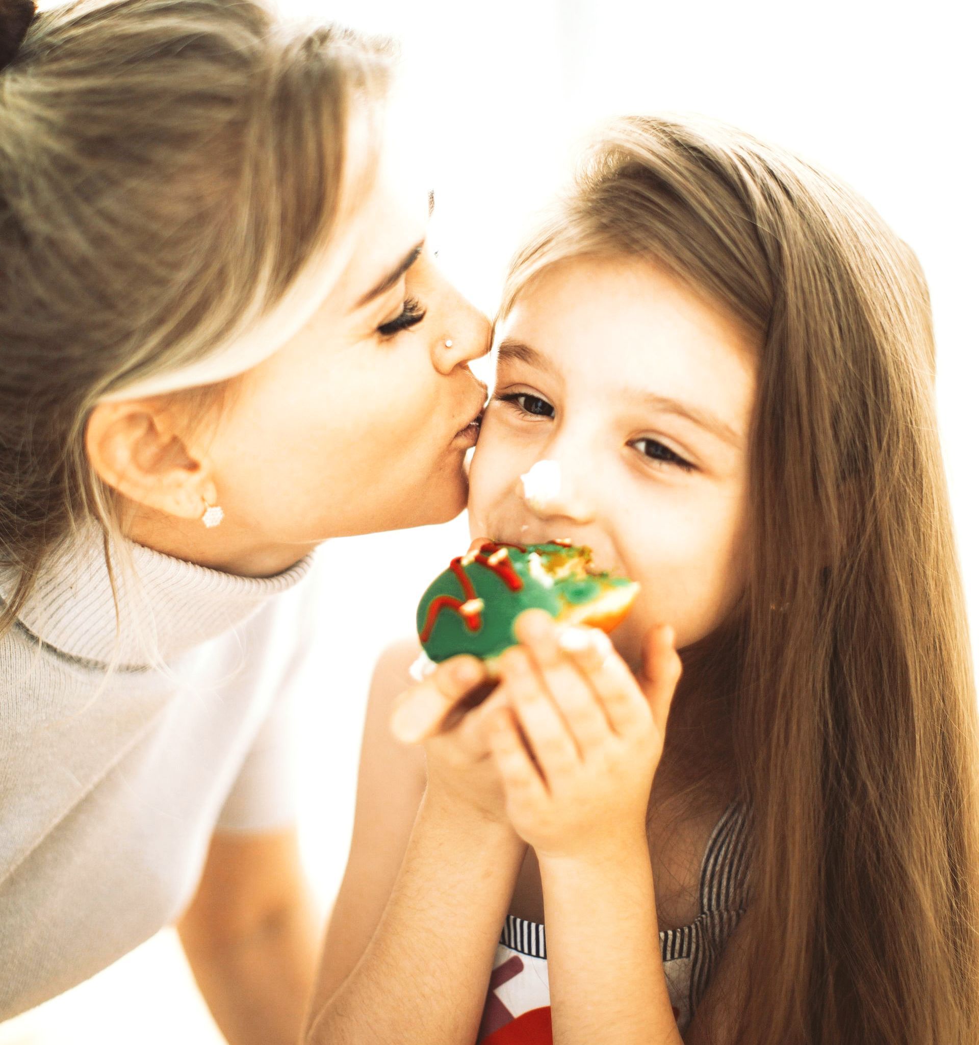 Eine elegant gekleidete Mutter küsst ihre 6jährige Tochter auf die Wange. Das Mädchen isst gerade ein Törtchen und hat die Nase mit Zucker verschmiert. Man kann die Liebe zwischen den beiden spüren.