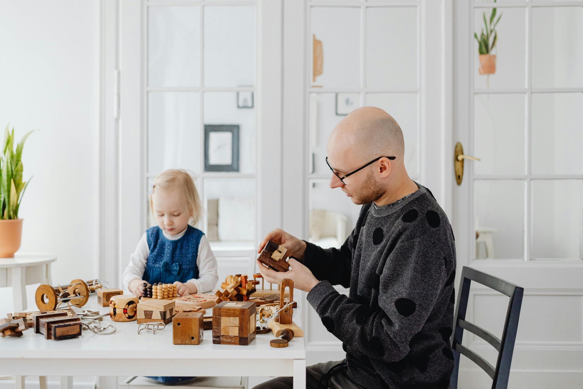 Ein Vater spielt mit seiner  4jährigen Tochter am Esstisch. Darauf befinden sich sehr viele Holzspielzeuge und Logikspiele. Der Raum ist freundlich hell und nüchtern.