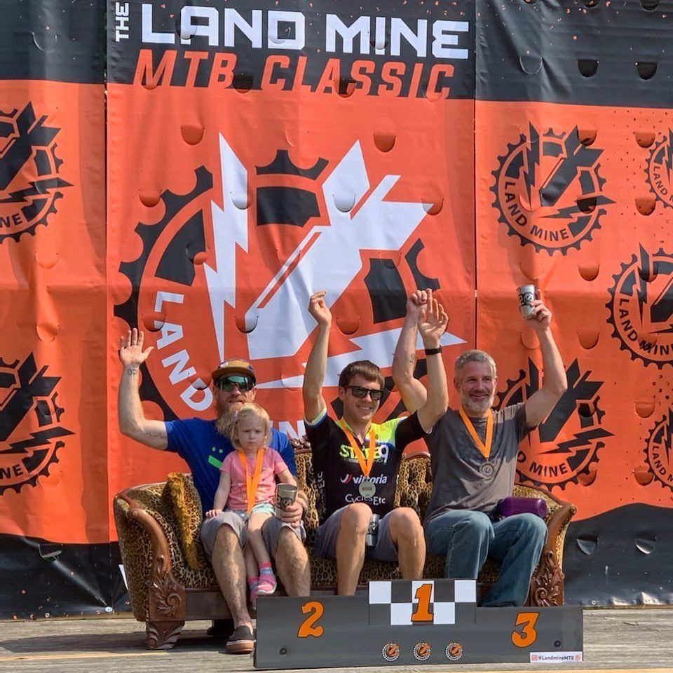 Four people on a podium waving at the Landmine MTB Classic event, with orange event banners behind them.