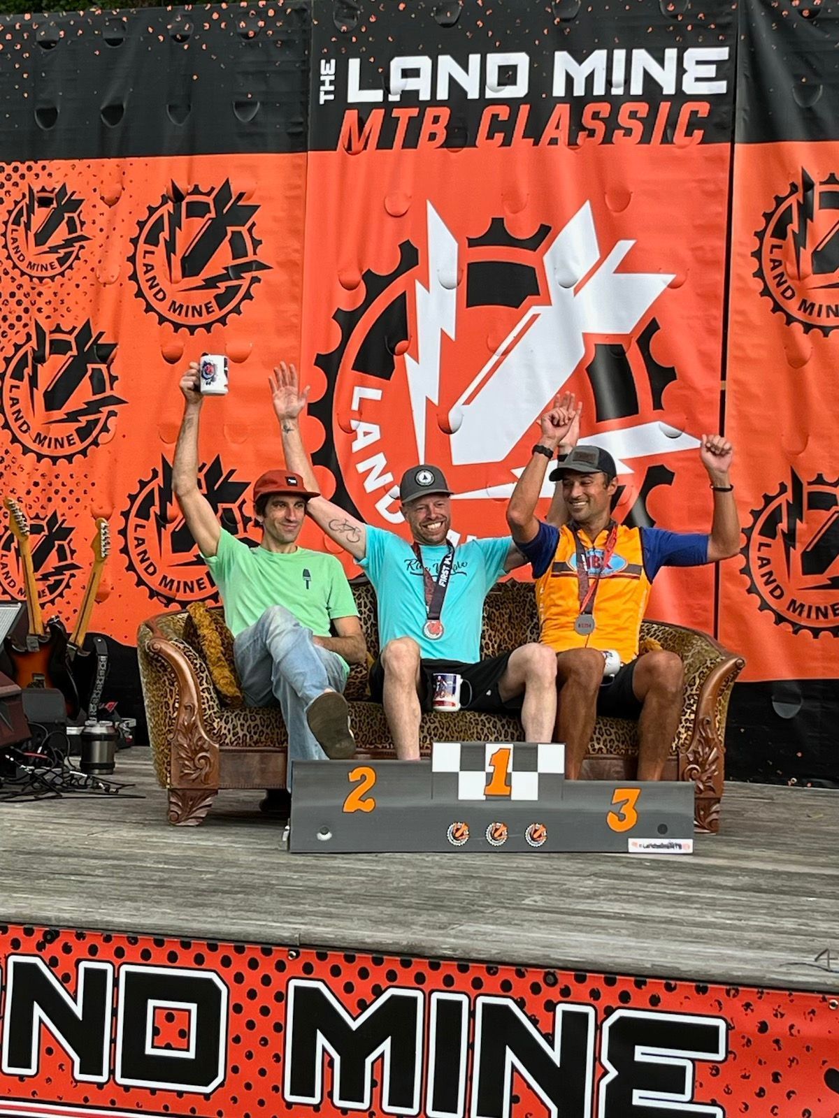 Three cyclists on a podium at Land Mine MTB Classic, raising trophies before a No Mine banner.