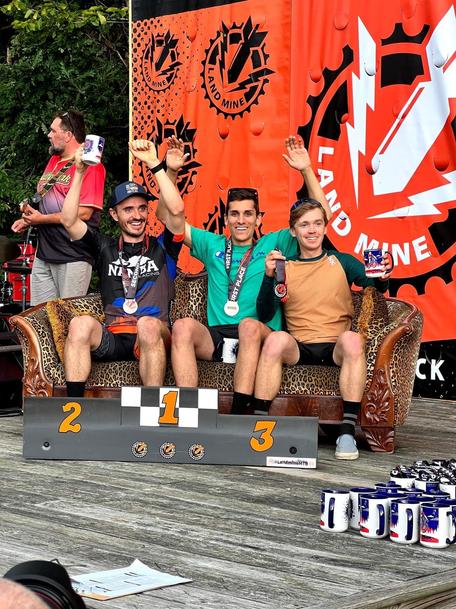 Three cyclists on a podium with trophies and medals at a cycling event, orange backdrop behind them.