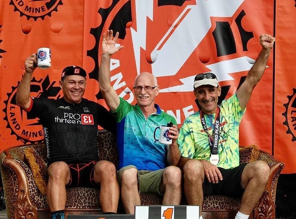 Three cyclists pose on a couch, smiling and raising their arms at a finish-line backdrop.