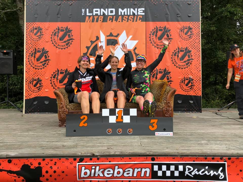 Three mountain bikers on a podium at Land Mine MTB Classic, celebrating with trophies and medals.