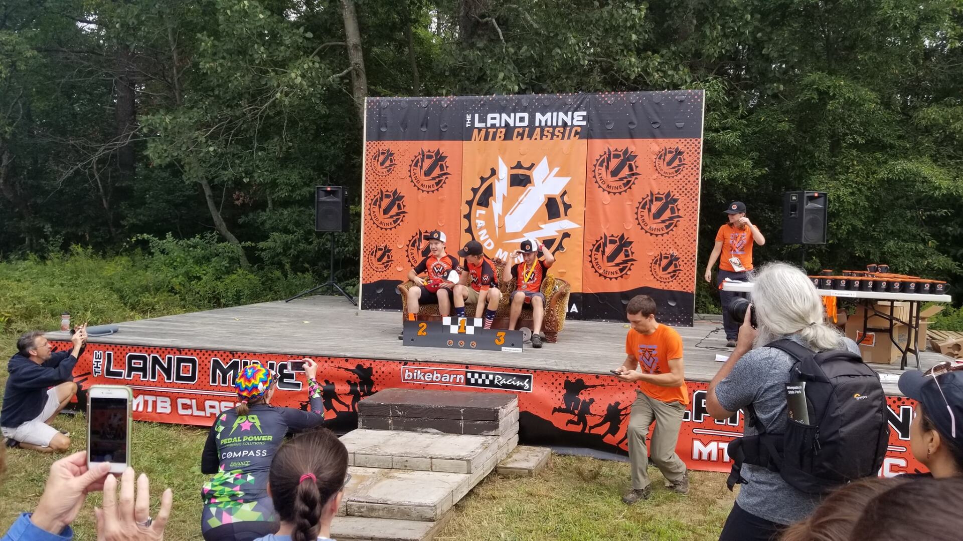 Outdoor cheer squad on an orange-and-black stage, with spectators watching from a grassy area.