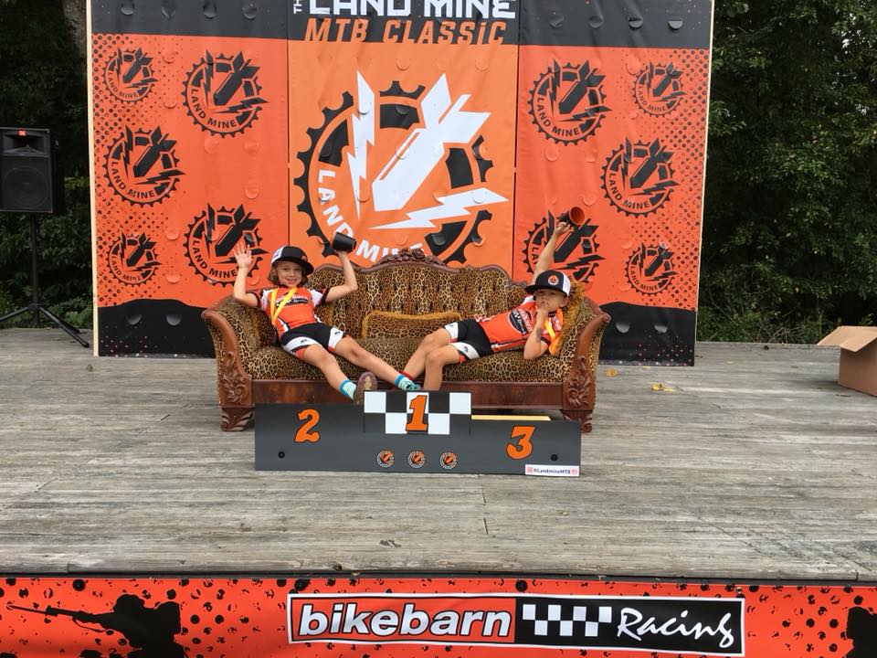 Two people relax on a podium at a bike race, with orange “Mtb Classic” and “bikebarn racing” banners behind them.