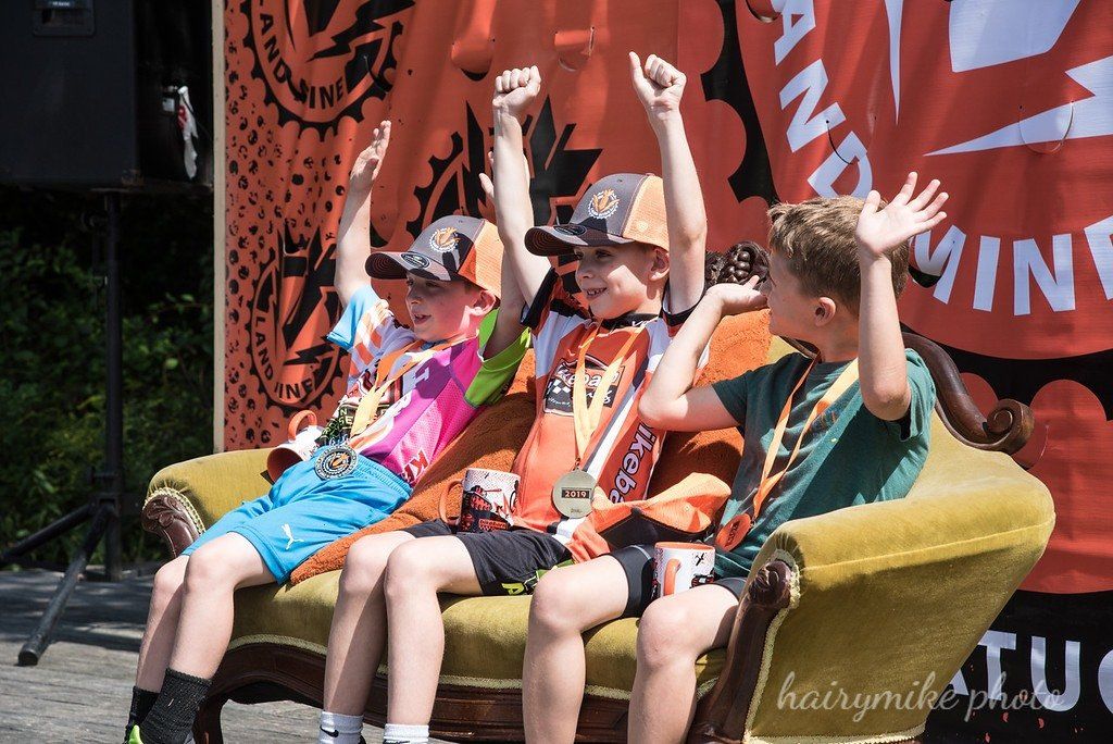 Three people in colorful outfits raise their arms while seated on a yellow couch at an outdoor event