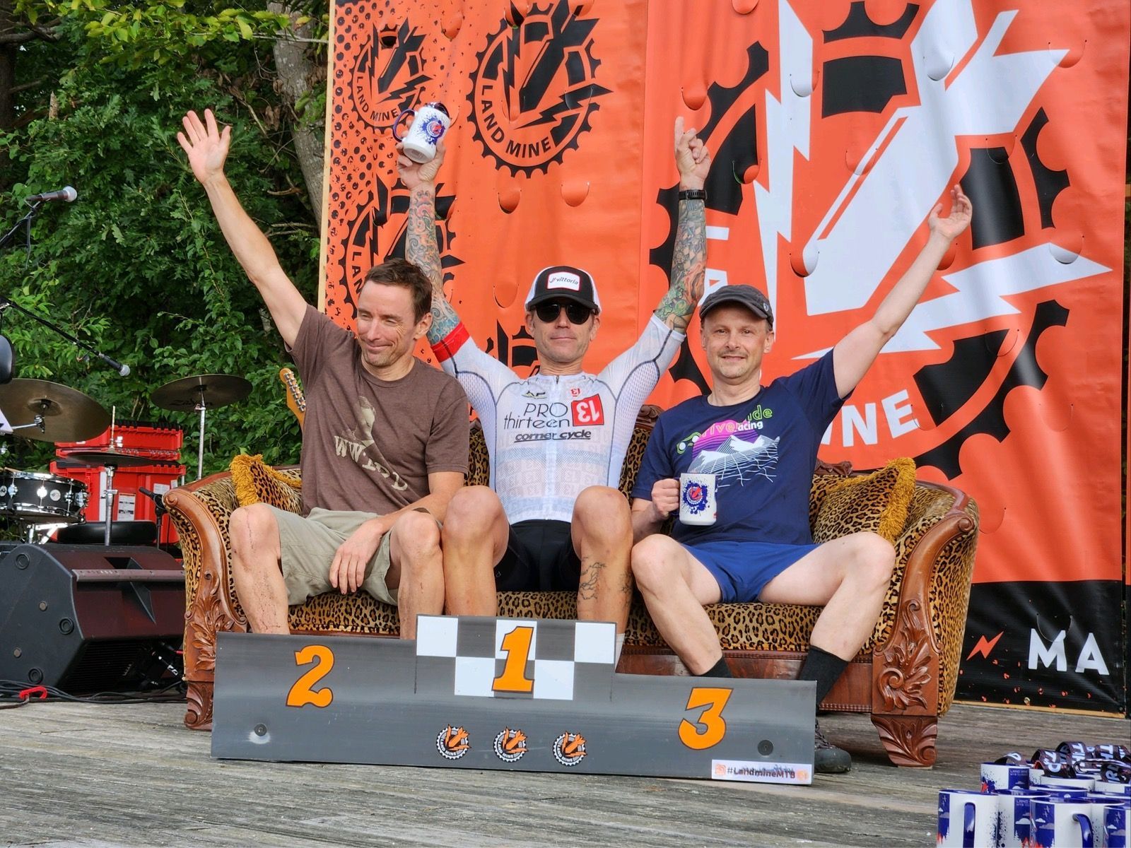 Three people on a winners' podium at a mountain bike race, arms raised before an orange backdrop.
