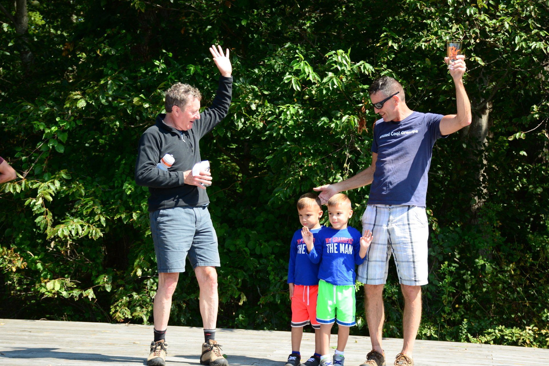 Two adults and two children waving outdoors in a sunny park-like setting