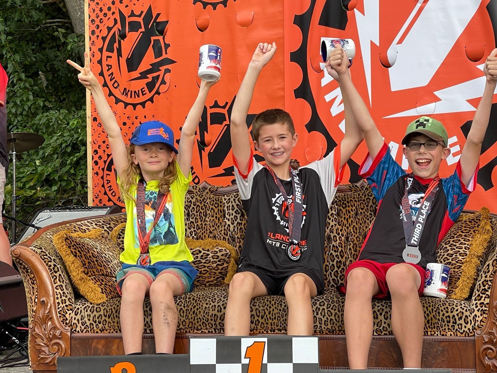 Three children in medals sit on a bench and raise cans in celebration at an outdoor event.