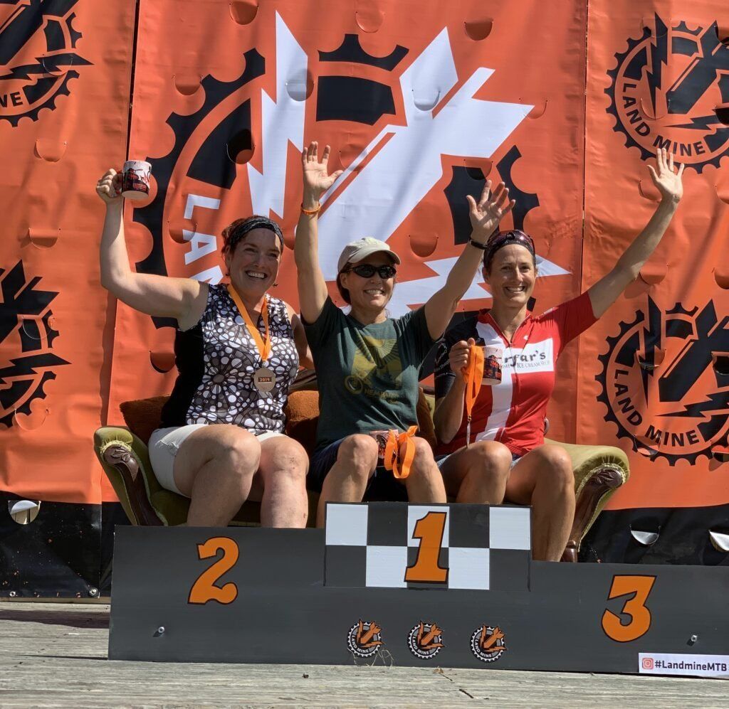 Three athletes on a winners’ podium raising arms, with orange race banners behind them.