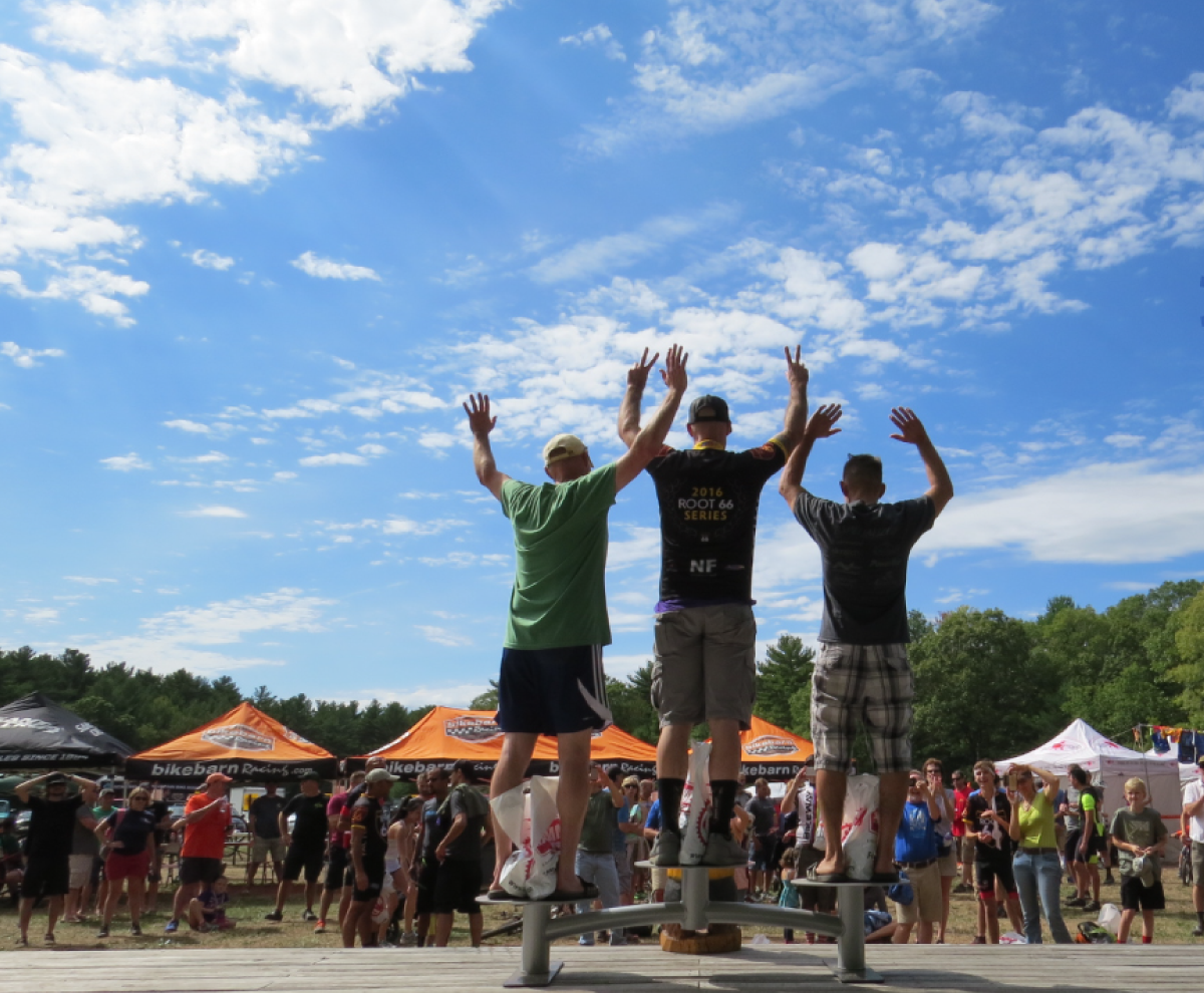 Three people on a platform cheering with raised arms at an outdoor festival under a blue sky