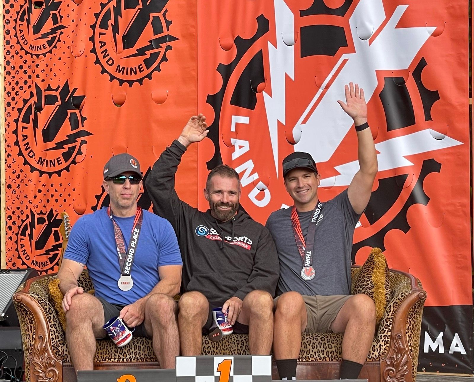 Three medalists sit on a podium, smiling and raising their arms before an orange race banner.