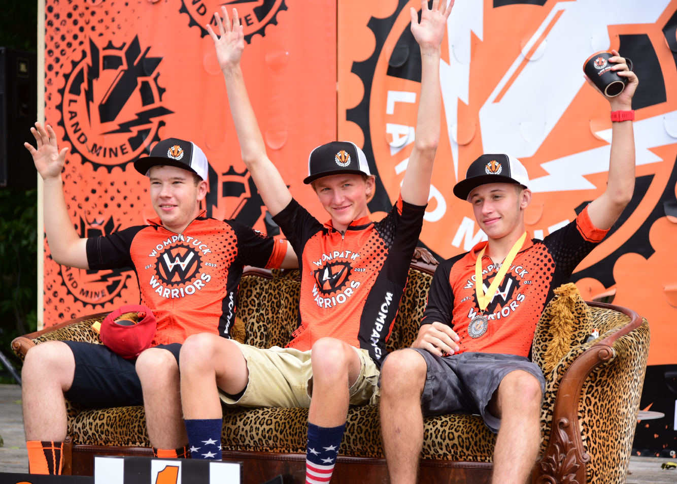 Three men in orange sports jerseys cheer on a bench at an outdoor event.