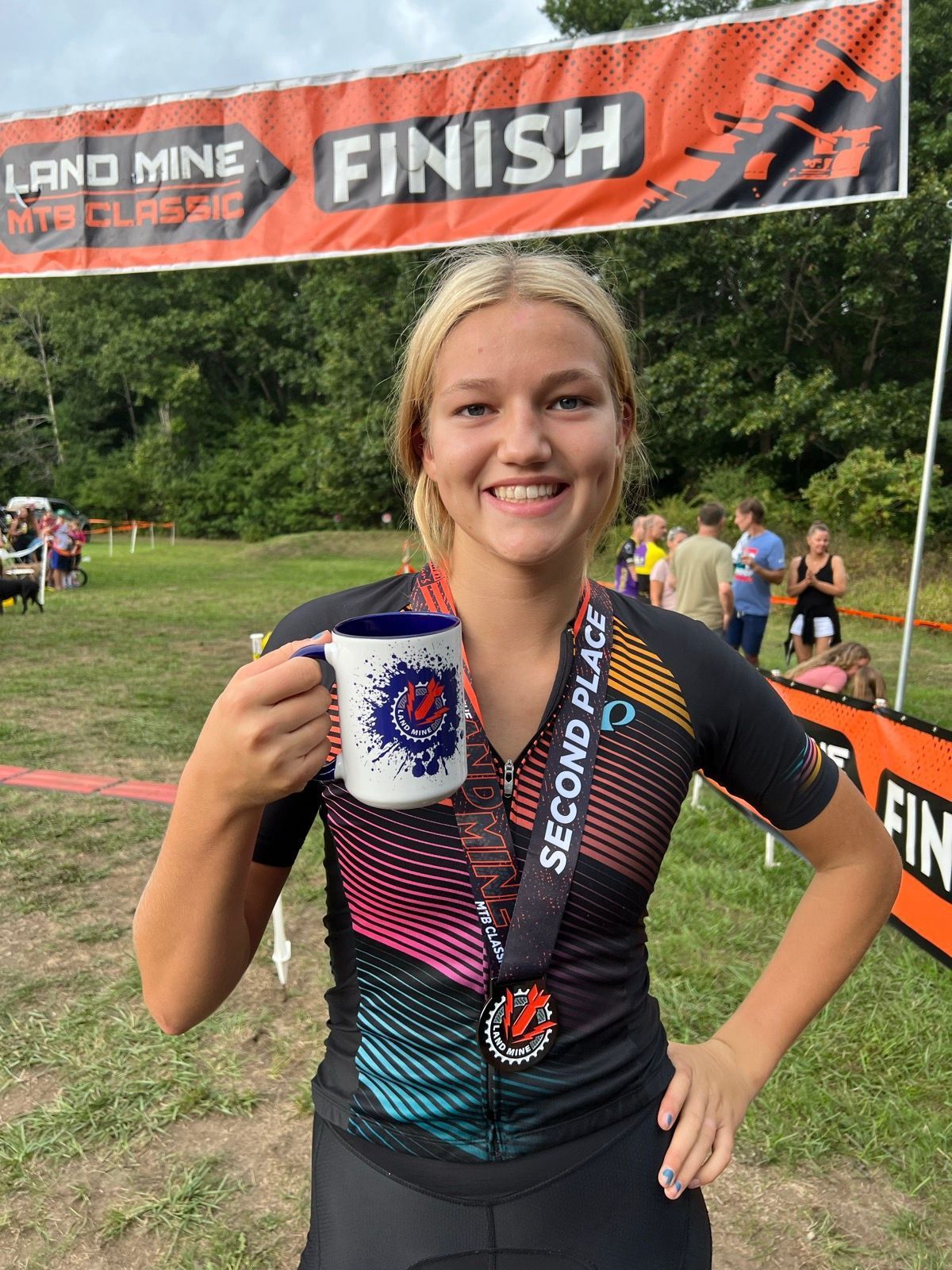 Smiling runner holding a mug at the finish line, wearing a black and teal race jersey.