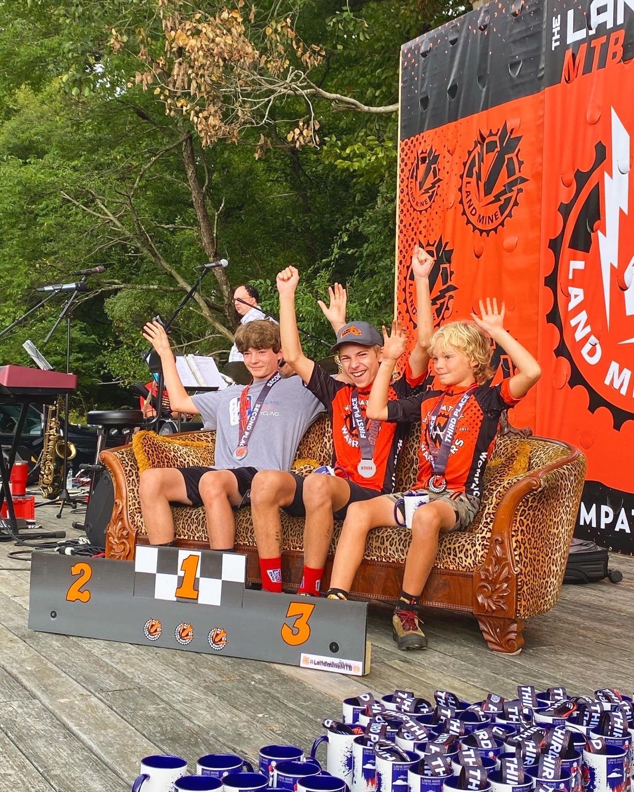 Three people on a winners' podium at an outdoor event, waving with orange and black decorations behind them.