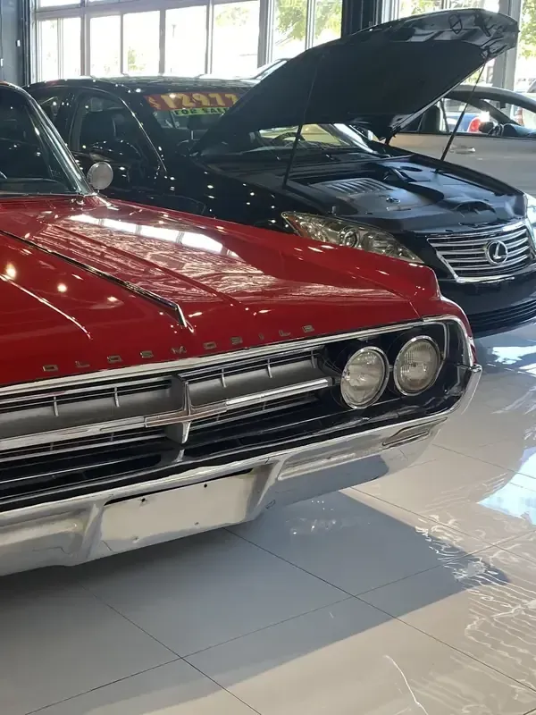 Red classic car in a showroom, with a black car behind it and its hood open.