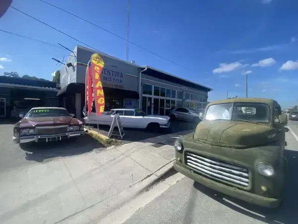 Vintage cars parked outside a storefront with a bright vertical sign under a blue sky