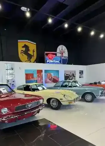 Classic car showroom with red, yellow, and silver vintage cars under brand signs and ceiling lights