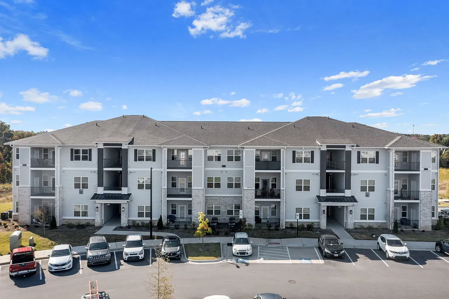 Exterior view of a modern three-story apartment building with balconies and a row of parked cars in front.