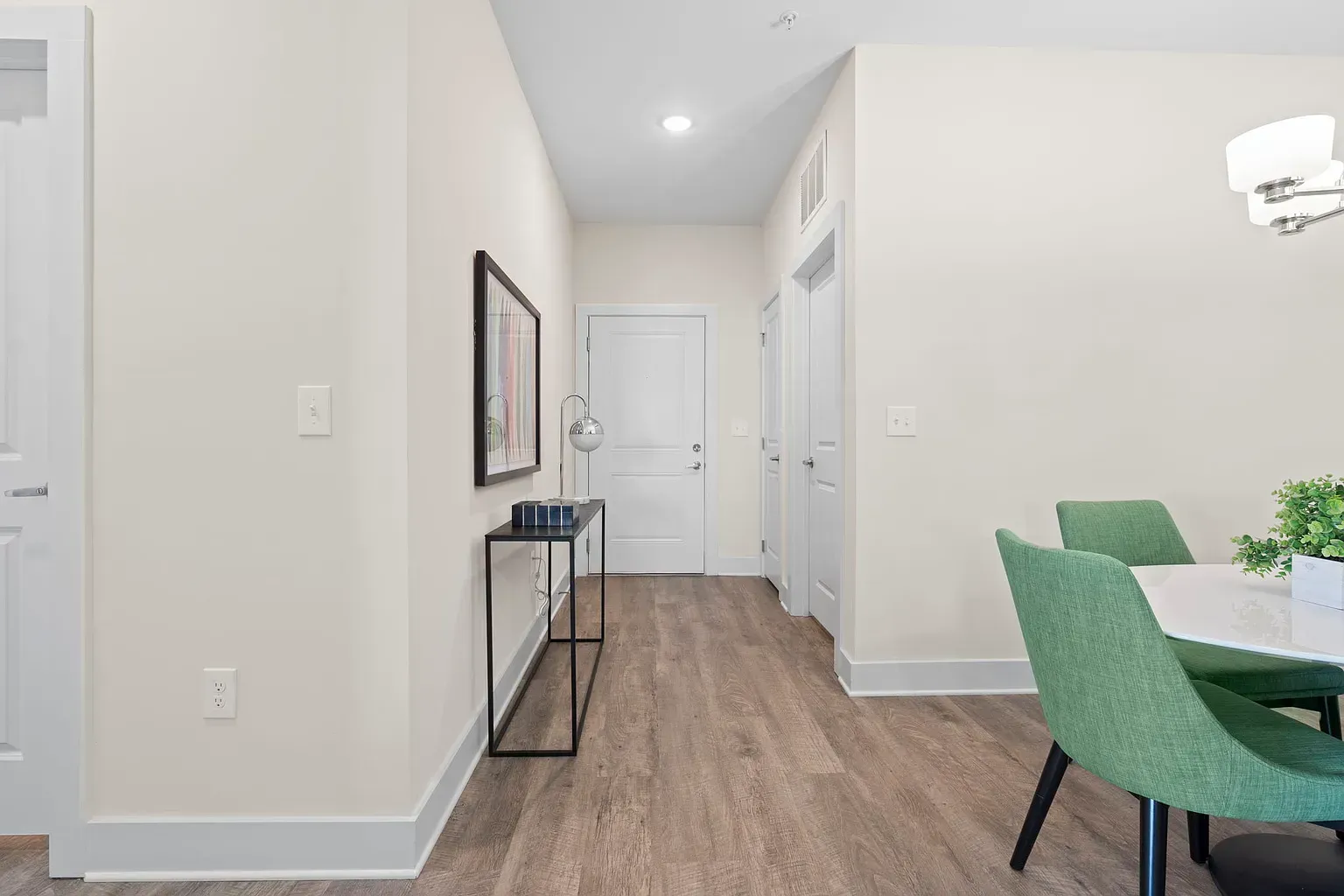 Interior apartment hallway with a console table, wall art, and a dining area with green chairs.