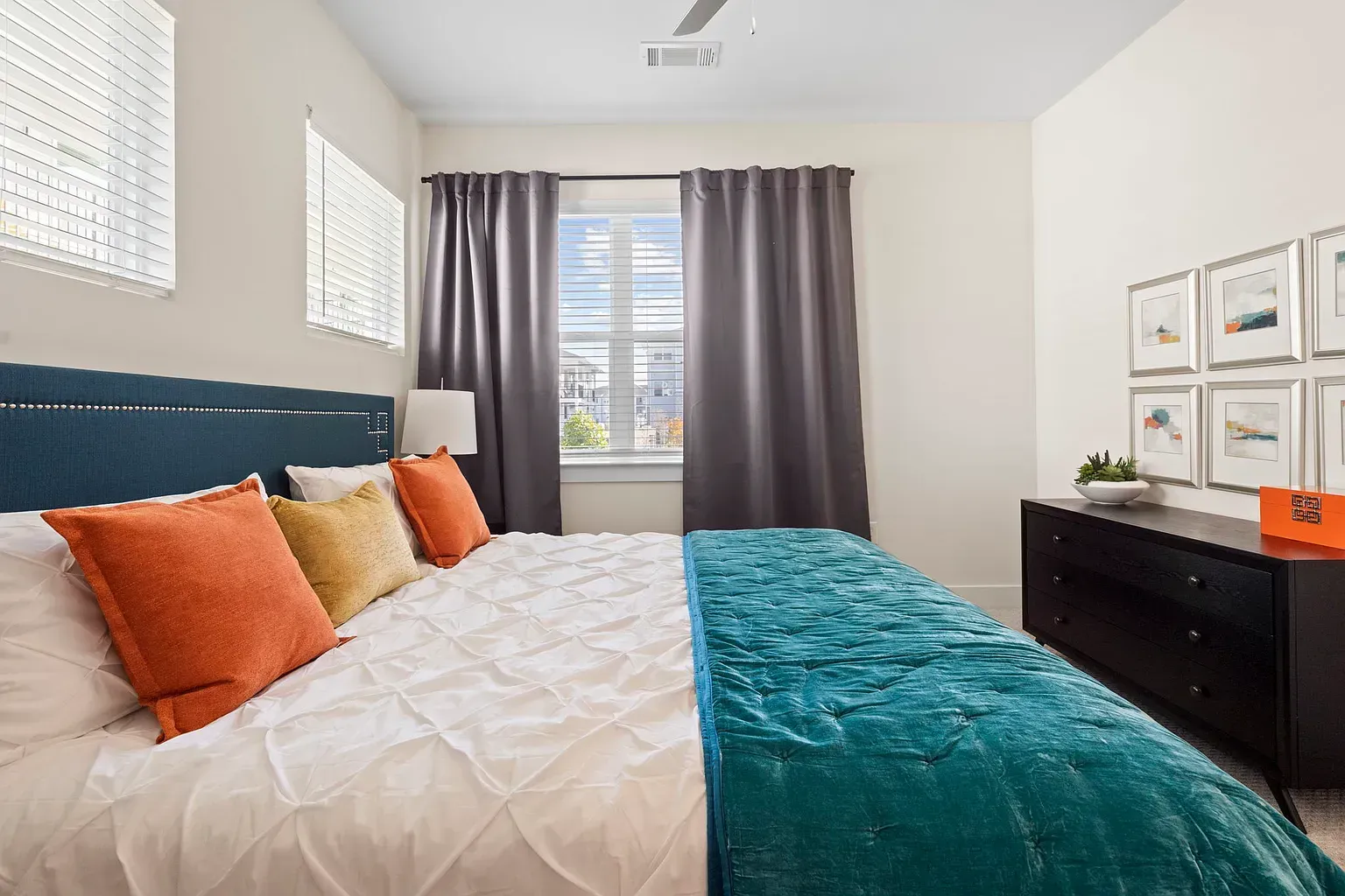 Bedroom in an apartment with a large bed, teal quilt, colorful pillows, and a dark dresser.