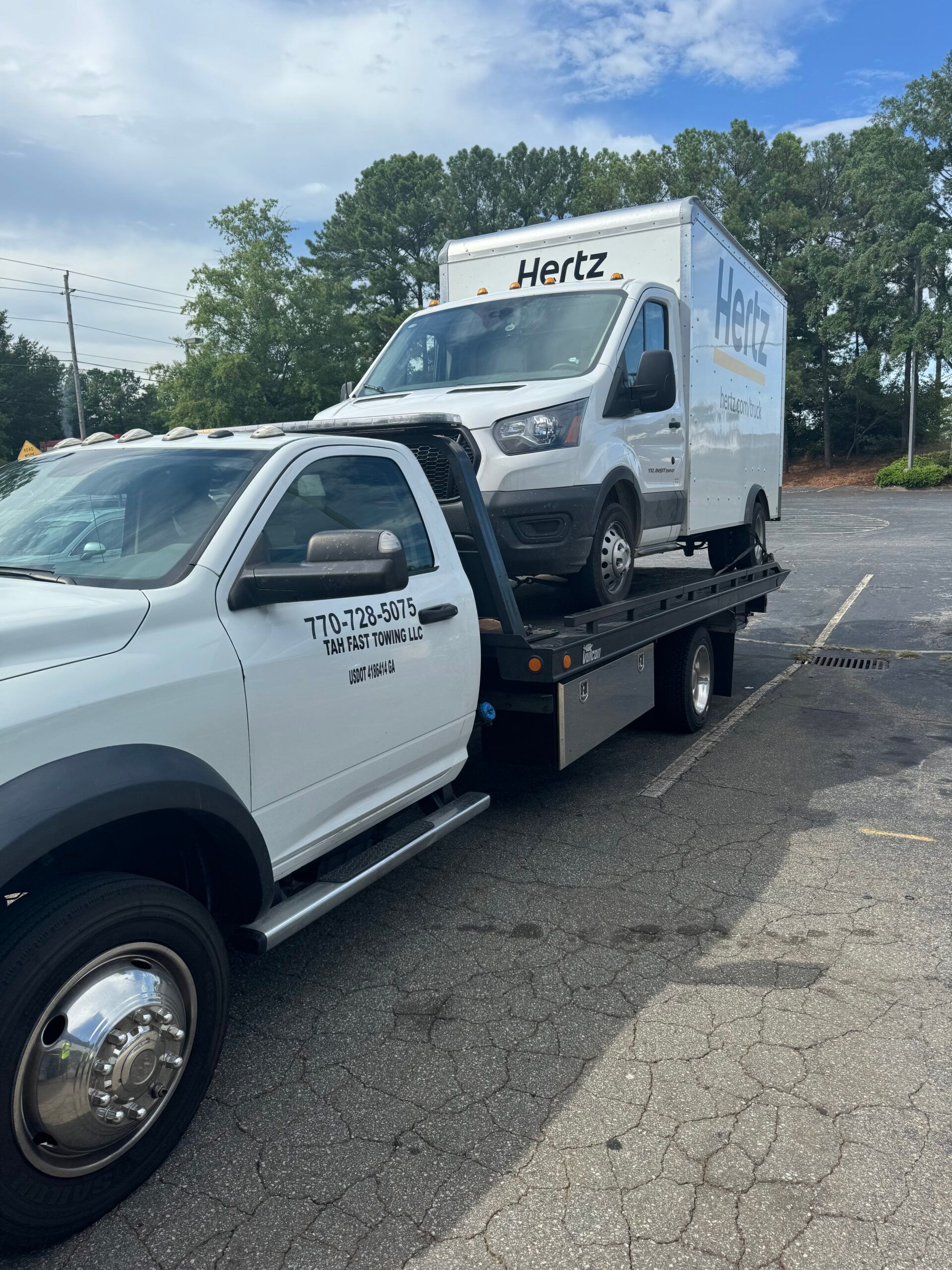 A tow truck is towing a hertz van in a parking lot.