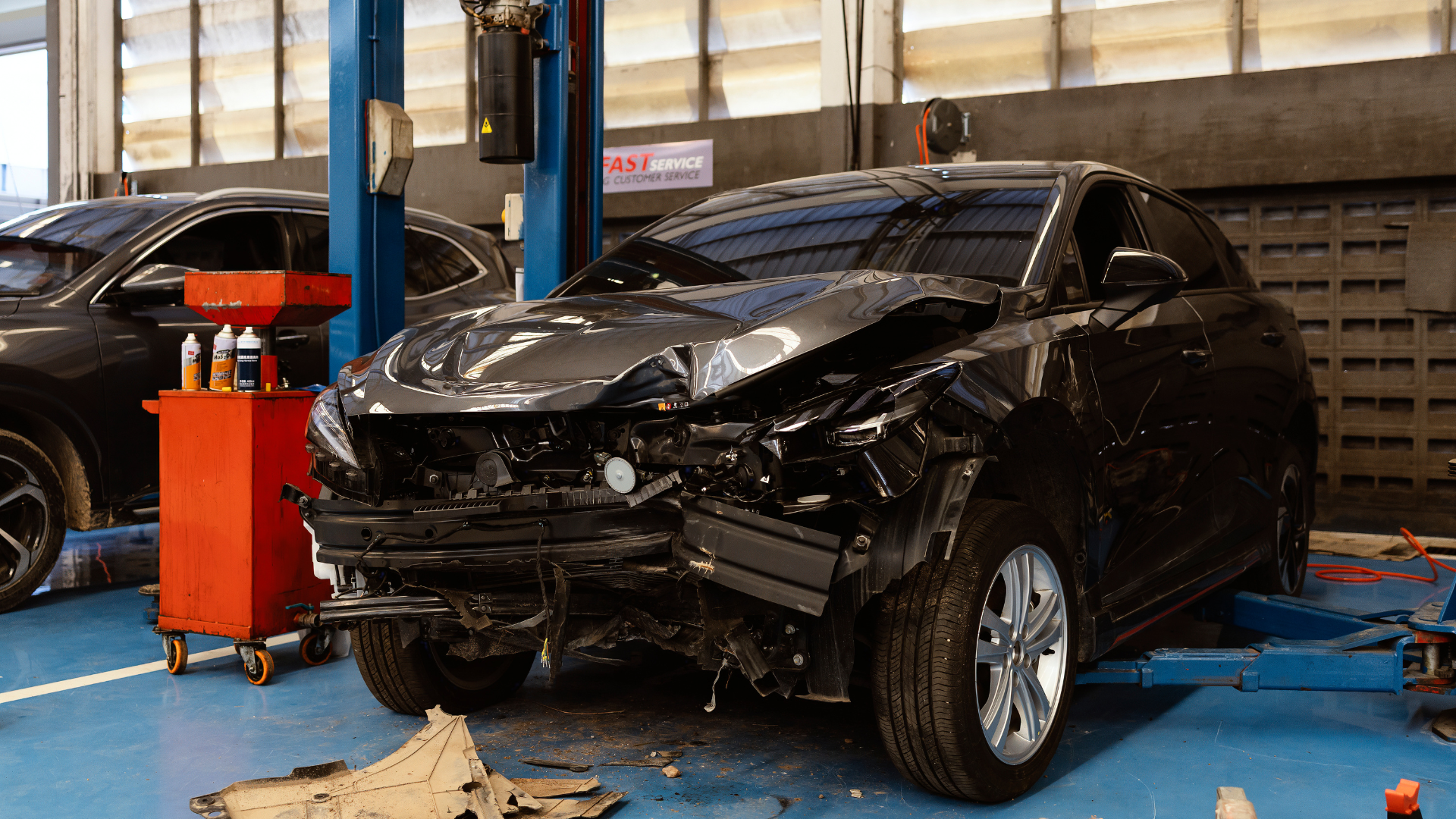 Damaged black car in a repair shop; front end heavily damaged. Other car and equipment in the background.