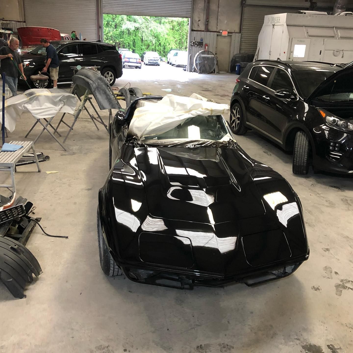 Black sports car in a body shop, covered with a white sheet. Other cars and people in the background.