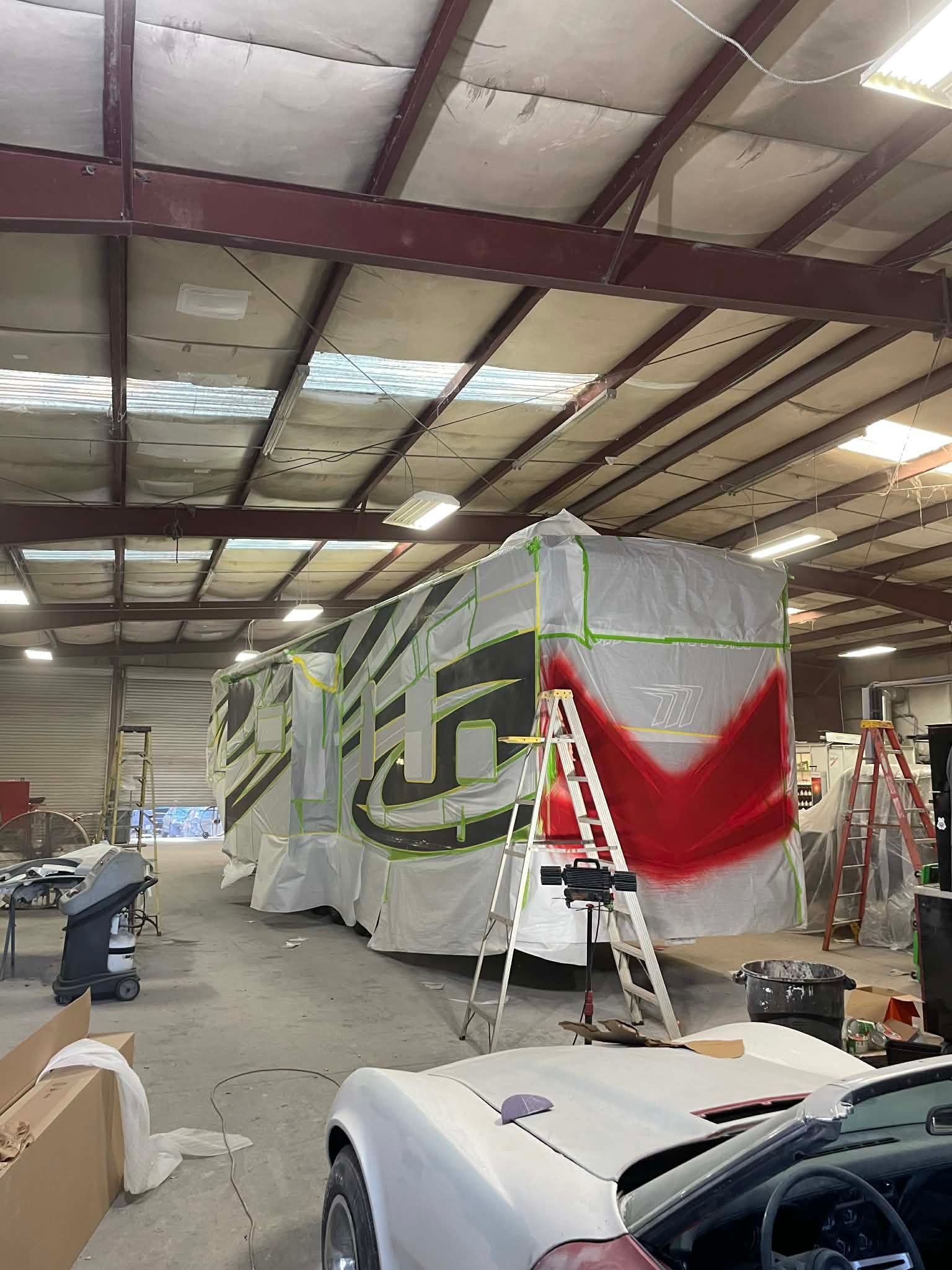 Large custom RV under construction in a warehouse, with scaffolding and a white car in the foreground.