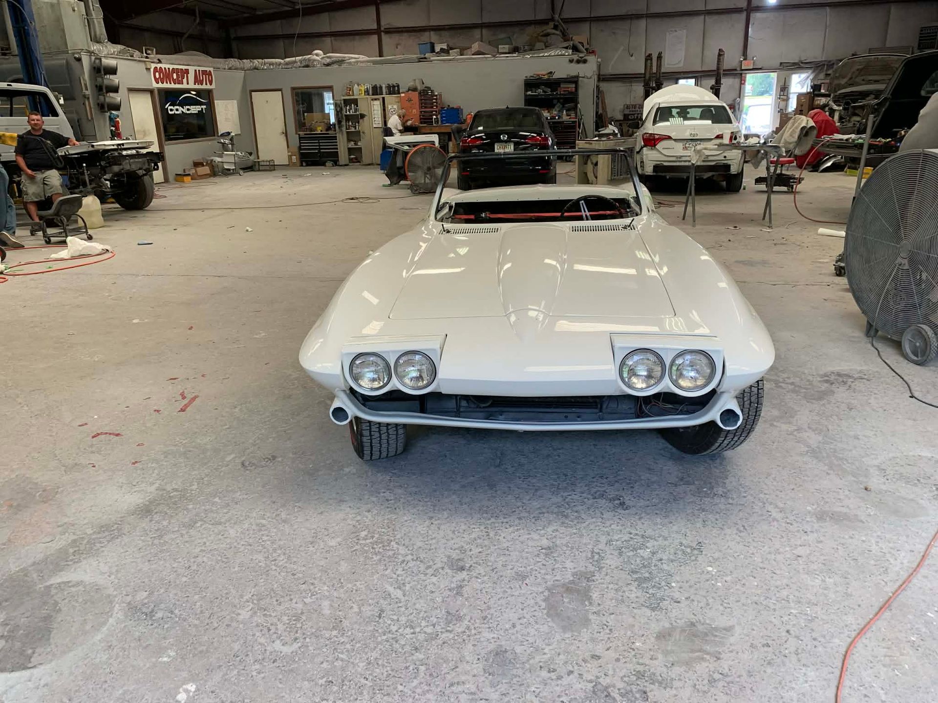 White classic convertible car in a workshop garage, front view with headlights on display.