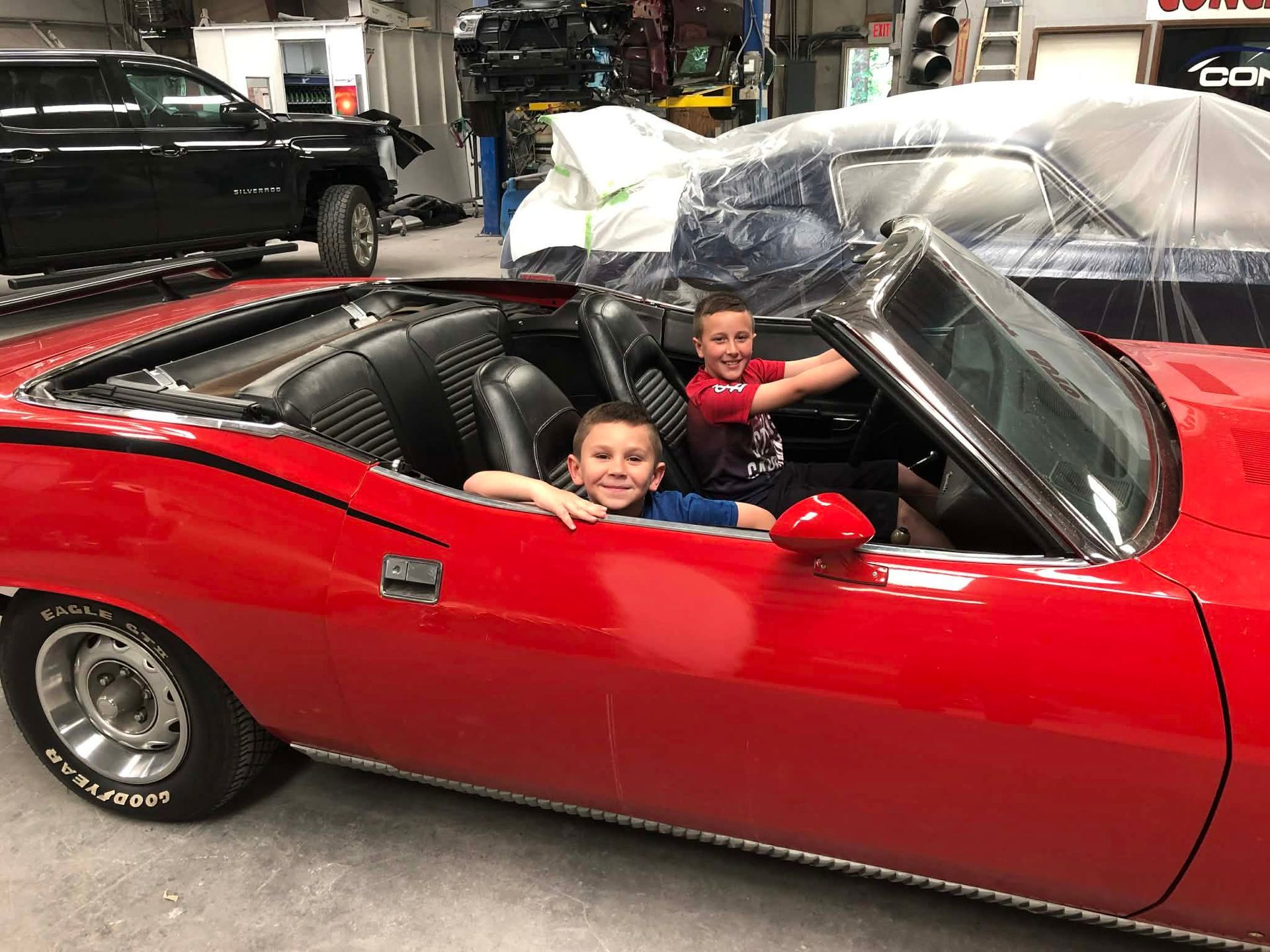 Two children smiling in a red convertible with a plastic-covered roof in a garage
