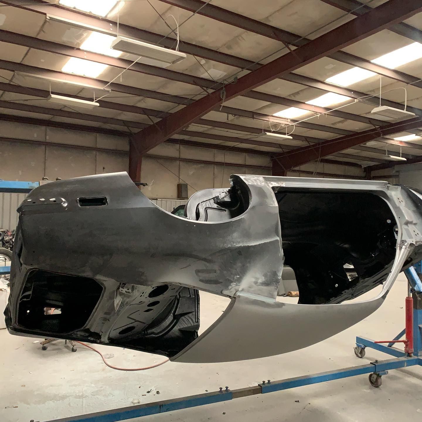 Car body in a workshop, grey primer, suspended on a blue frame, awaiting paint.
