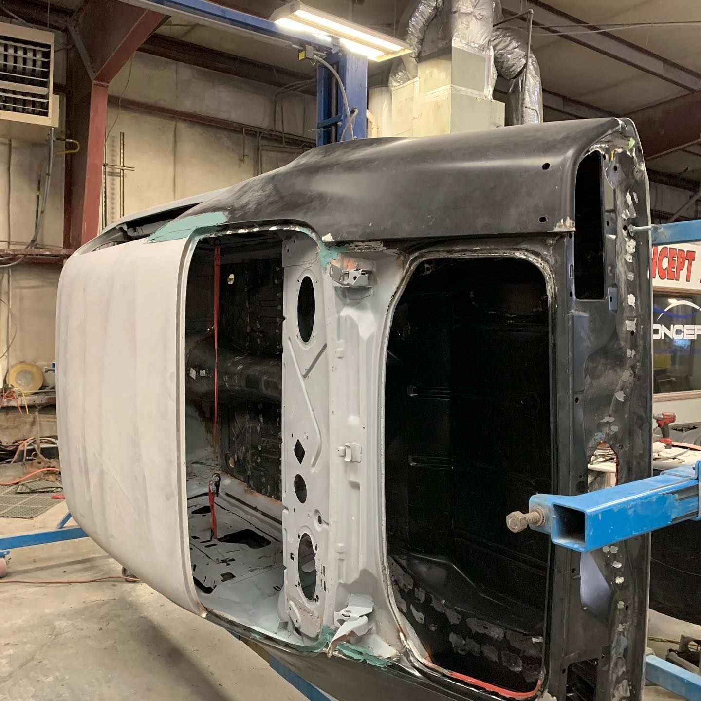Car body in a workshop, upside down for restoration. Black and gray colors, metal structure visible.