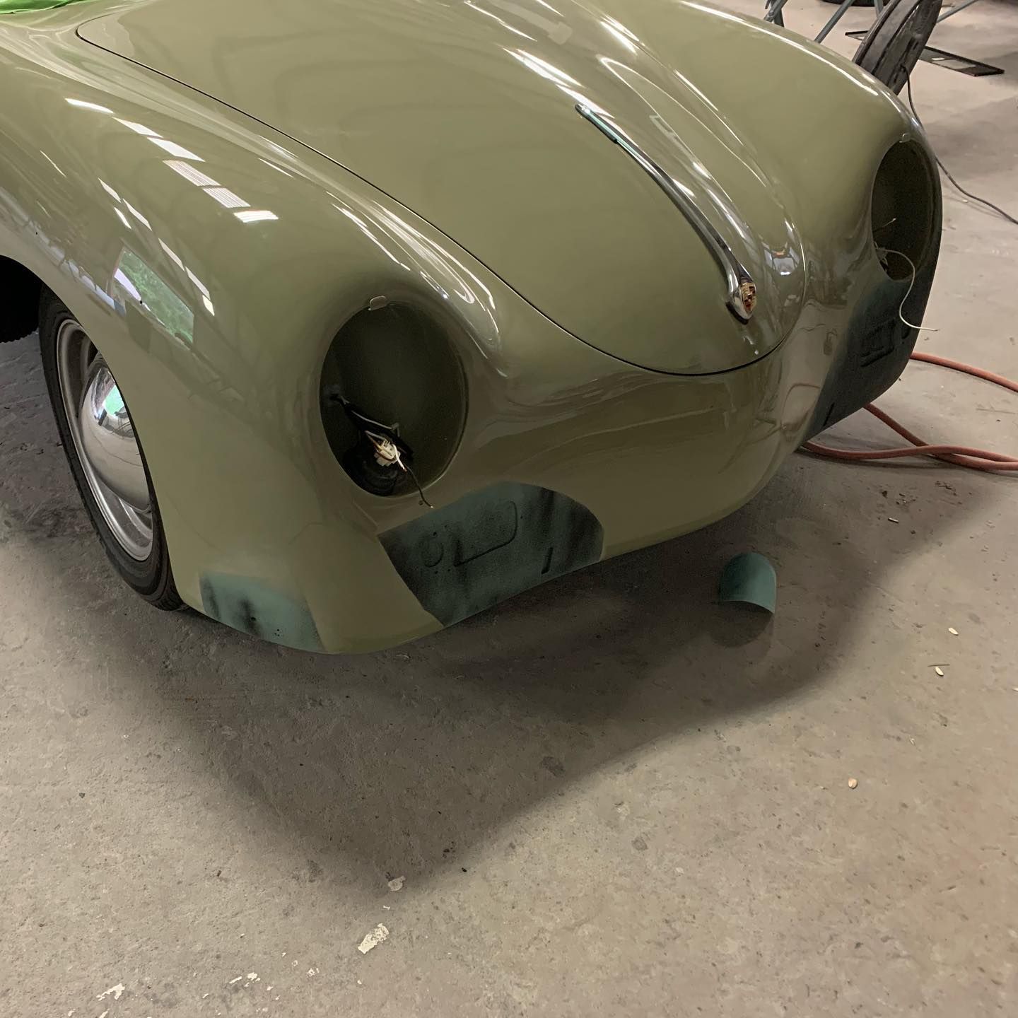 Front end of a classic green car in a repair shop. Light green paint, chrome accents, and dark primer visible.