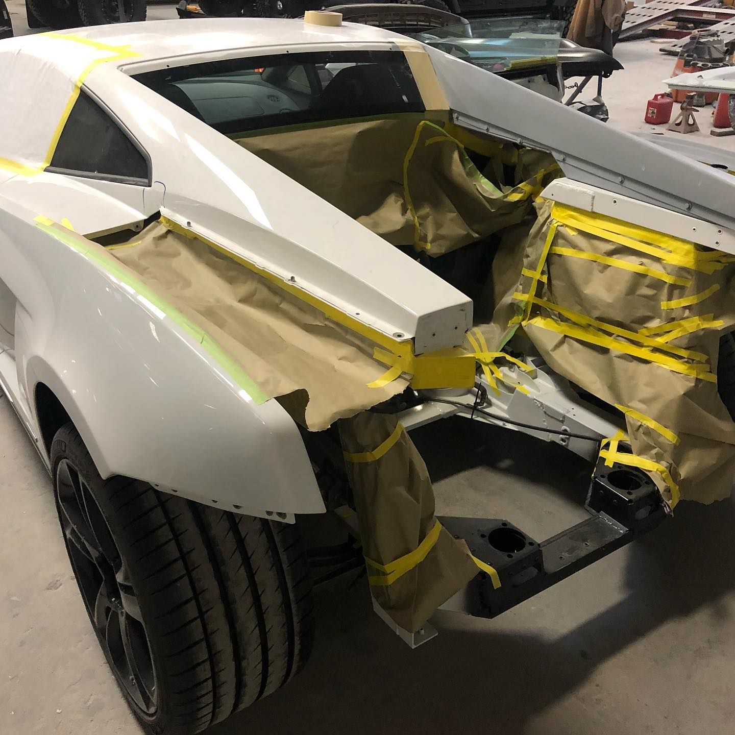 White car with rear section disassembled, masked with yellow tape and covered with brown paper, in a workshop.